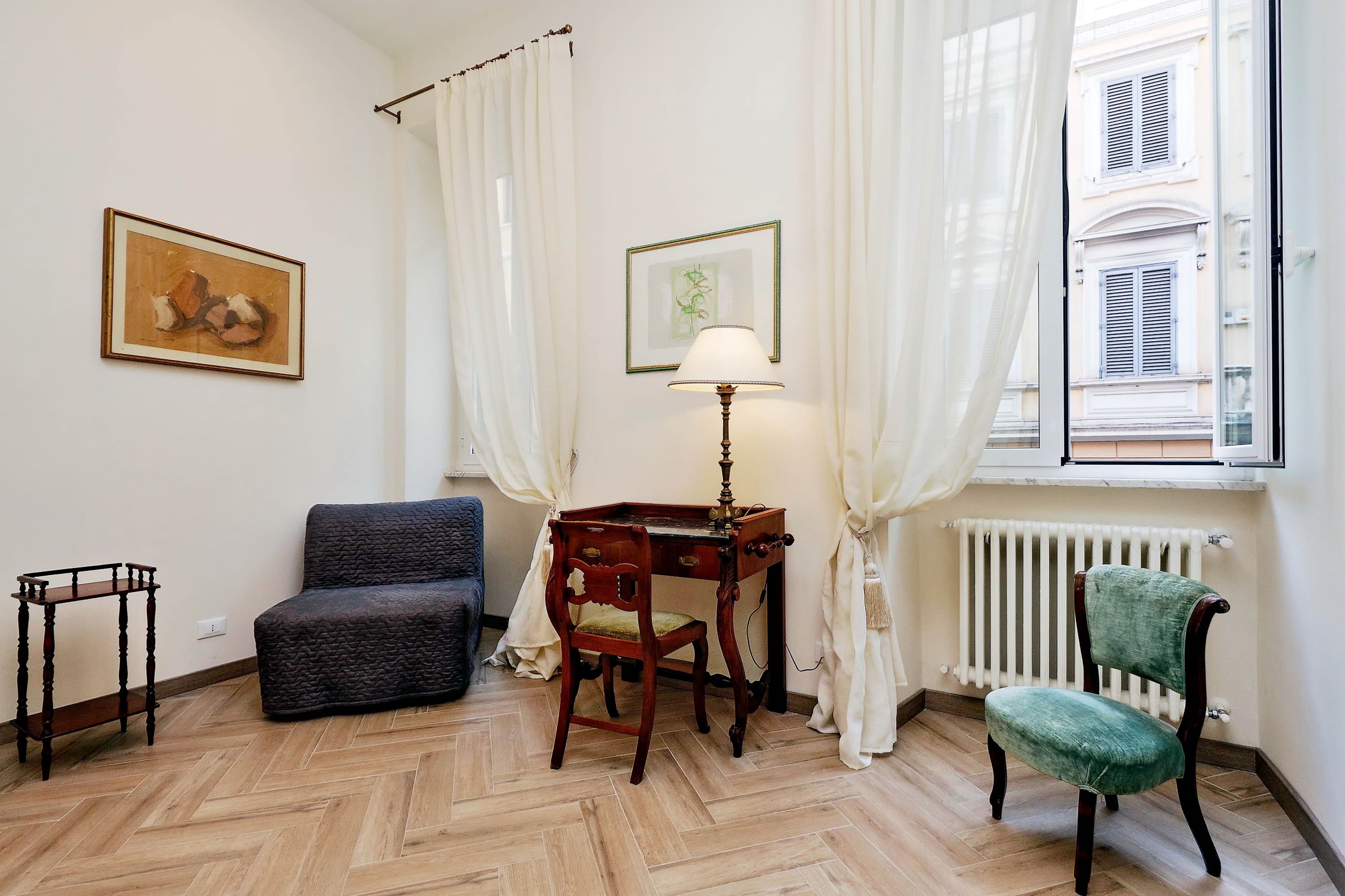 Una habitación con piso de espiga, un escritorio de madera con una lámpara, un sillón tapizado en verde, un sofá oscuro y dos cuadros en la pared.