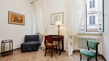 Una habitación con piso de espiga, un escritorio de madera con una lámpara, un sillón tapizado en verde, un sofá oscuro y dos cuadros en la pared.