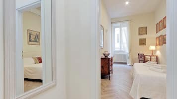 Habitación de hotel con un espejo grande, una cama con sábanas blancas, un aparador de madera, una mesita con lámpara y una ventana con cortinas.