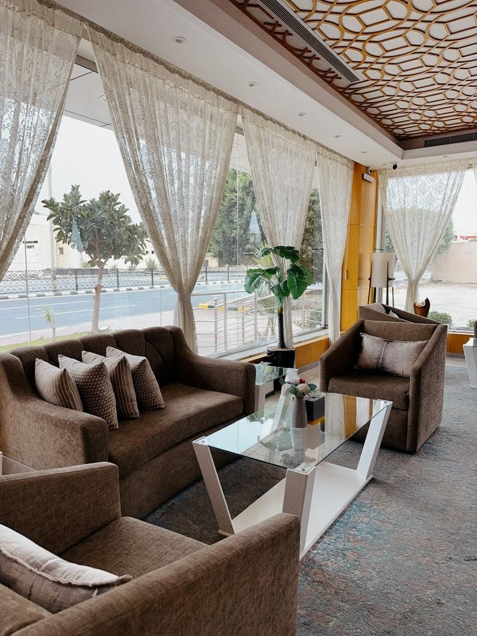 A modern hotel room with a sofa set, a glass coffee table, and a view of the street through large windows.