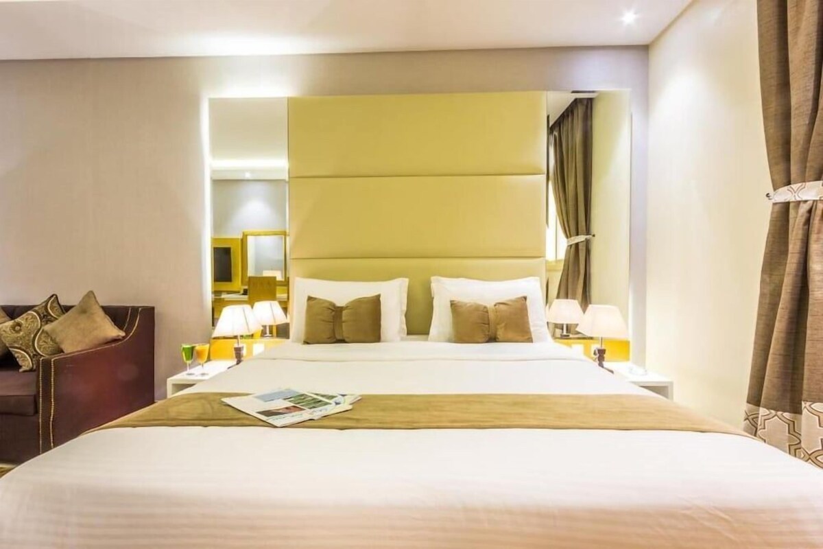 Habitación de hotel con una cama grande, un sofá, dos mesitas de noche con lámparas y un espejo.