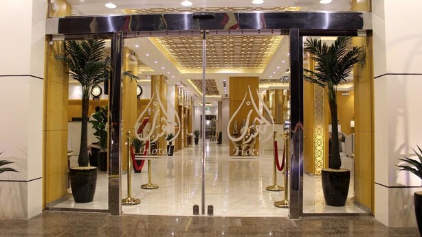 Entrada de hotel con puerta de vidrio, cuerdas de terciopelo rojo y plantas en macetas.