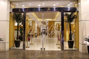 Entrada de hotel con puerta de vidrio, cuerdas de terciopelo rojo y plantas en macetas.