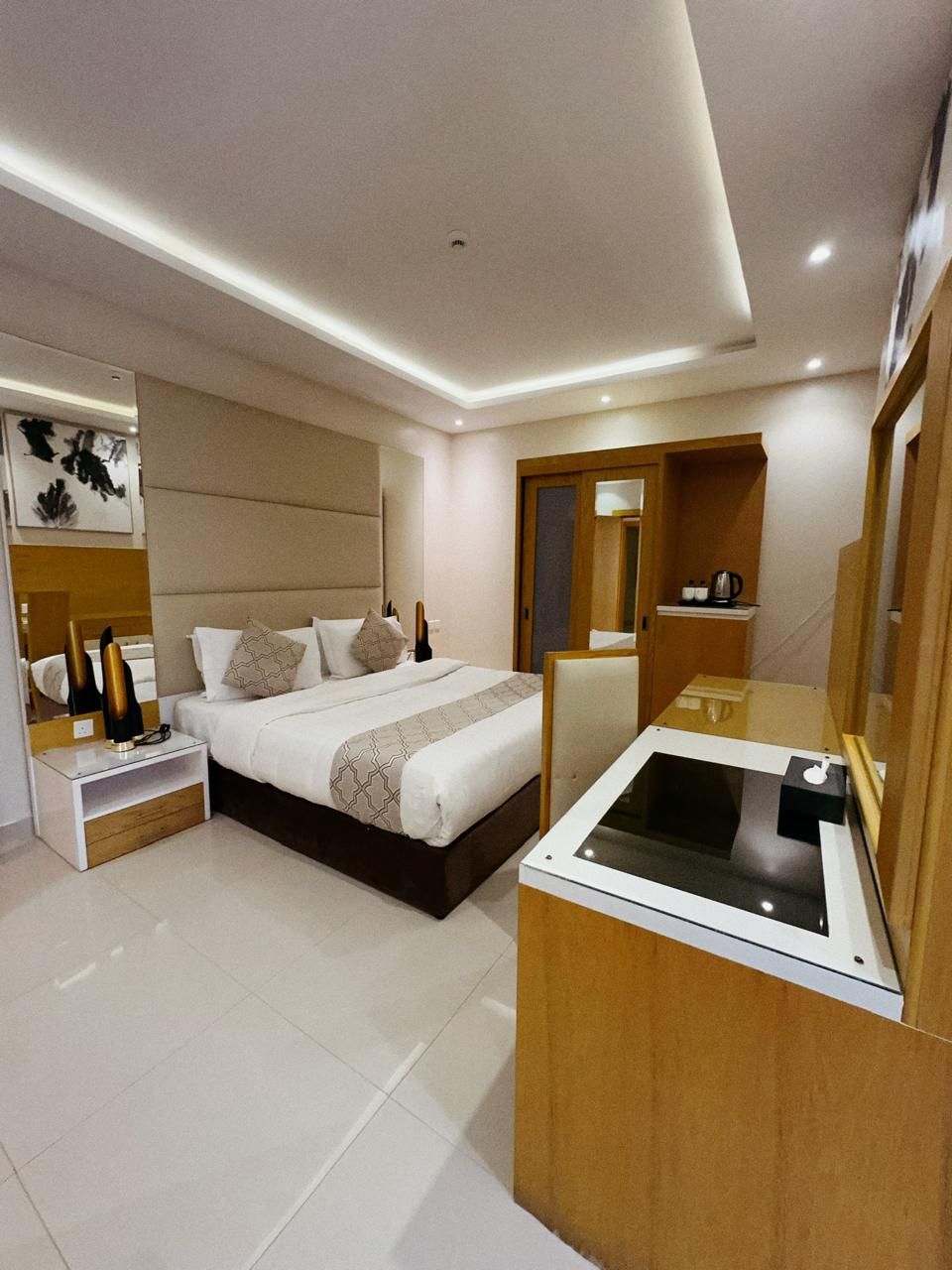 A modern hotel room with a large bed, a bedside table, a built-in desk with a mirror, and a small seating area.