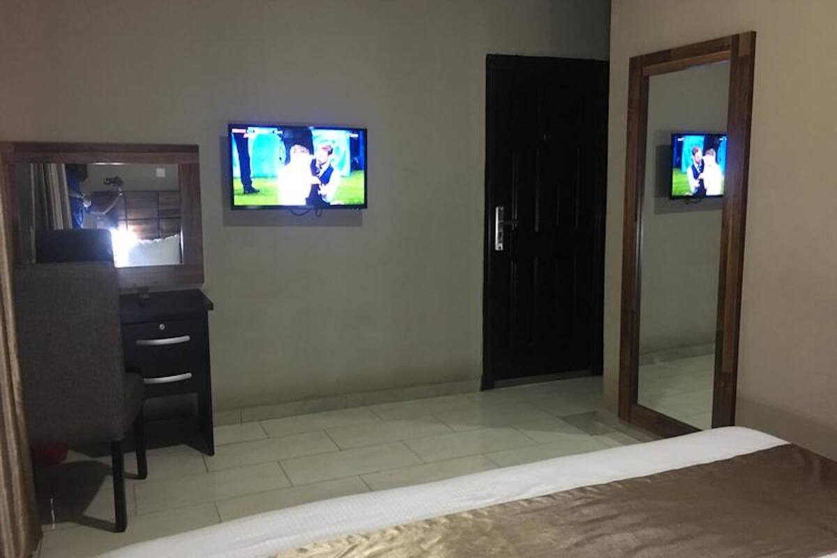 A hotel room with a bed, a desk, two televisions, a door, and a mirror.