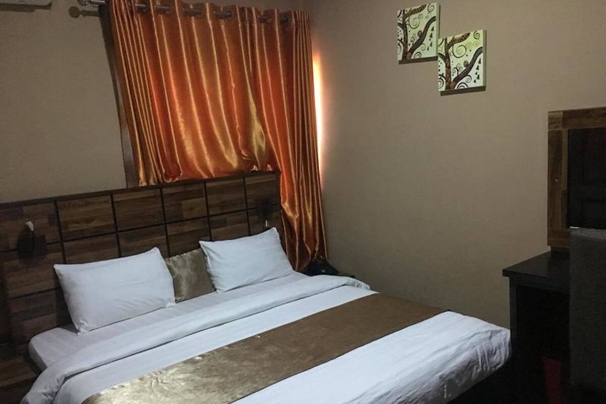A bed with white bedding and a brown accent, a wooden headboard, and a curtain.
