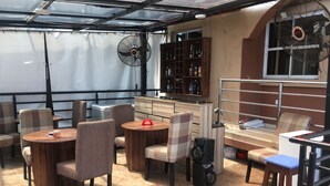 Un área de estar interior acogedora con mesas y sillas redondas, una zona de bar con estantes llenos de botellas, y dos ventiladores de techo.