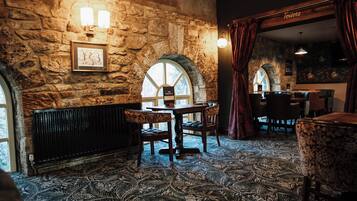 Una habitación con paredes de piedra, alfombra estampada, una mesa con sillas y un cartel que indica la ubicación de los baños.
