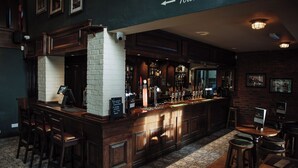 Un espace bar avec un mur en briques, un comptoir en bois et des tabourets. Une pancarte indique la direction des toilettes.