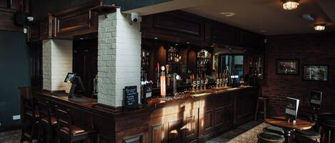 A bar area with a brick wall, wooden counter, and stools. There is a sign indicating the direction to the toilets.