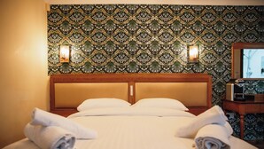 Un lit bien fait, avec des serviettes pliées, une tête de lit en bois et un papier peint à motifs, agrémenté d’appliques murales.