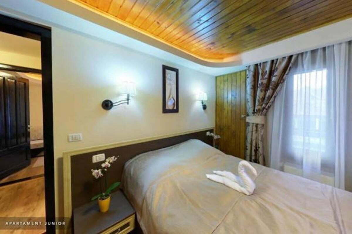 A hotel room with a wooden ceiling, a bed with a beige bedspread, two wall-mounted lights, a framed picture, and a window with curtains.
