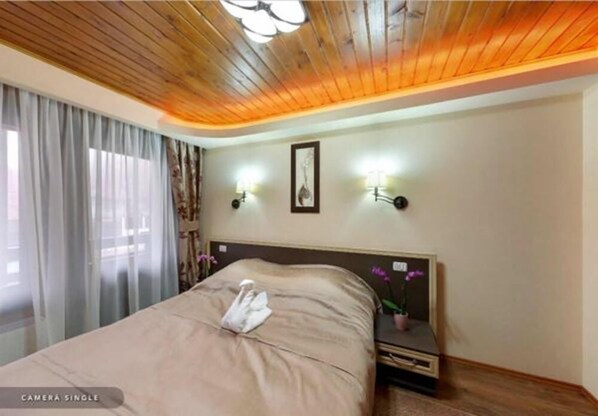 Habitación de hotel con techo de madera, cama con toalla, dos lámparas de pared y mesita de noche con planta.