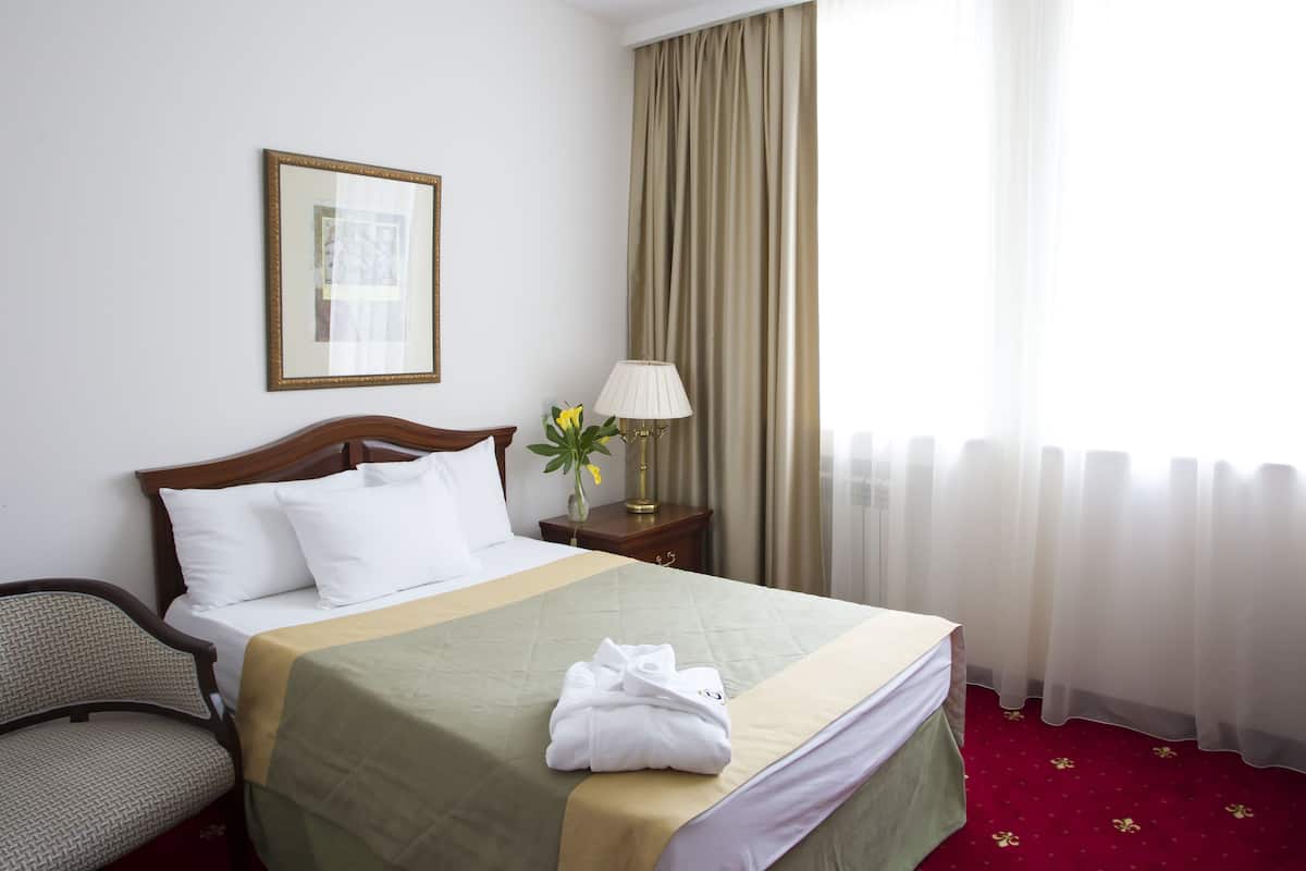 A neatly made bed with a green bedspread, white pillows, and a folded white towel on the bed. There is a wooden nightstand with a lamp and a vase with flowers beside the bed. A chair is visible to the left of the bed.