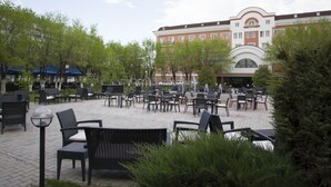 Un espace extérieur aménagé en terrasse, avec plusieurs tables et chaises, entouré de verdure et avec un bâtiment en arrière-plan.