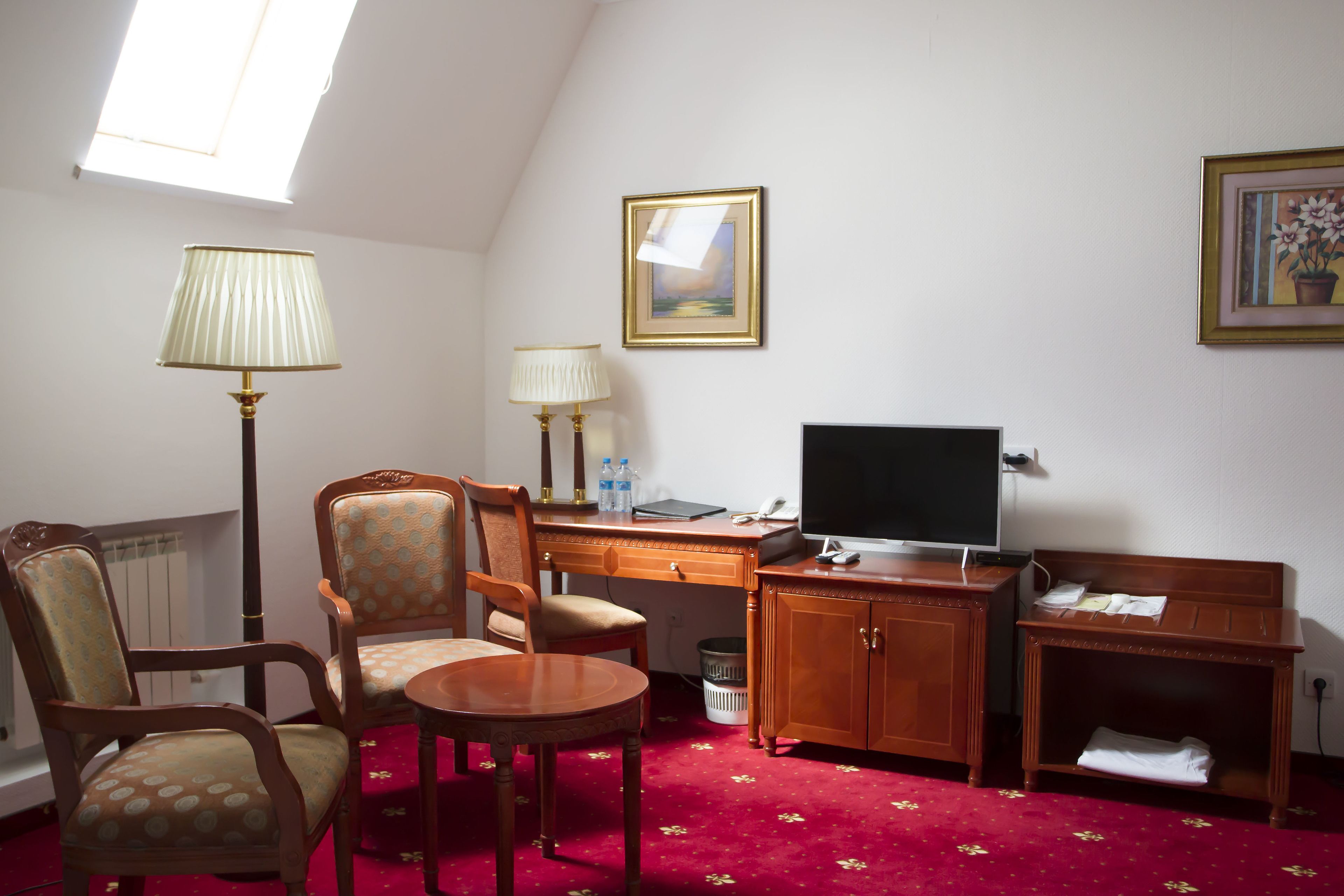 Ein Hotelzimmer mit Schreibtisch, zwei Stühlen, einem Fernseher, einer Lampe und einem roten Teppich.