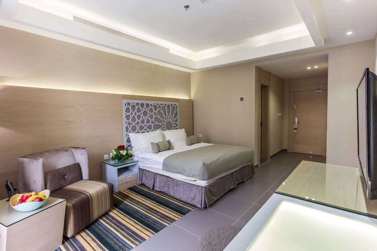 A modern hotel room with a large bed, a bedside table with a lamp, a chair, and a glass-top table.