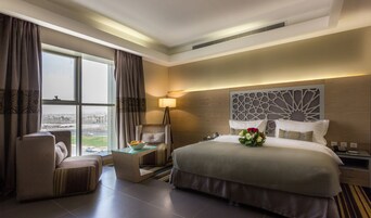 Une chambre d’hôtel avec un grand lit, un coin salon comprenant un fauteuil et une petite table, une table de chevet avec une lampe, et une vue sur la ville par la fenêtre.