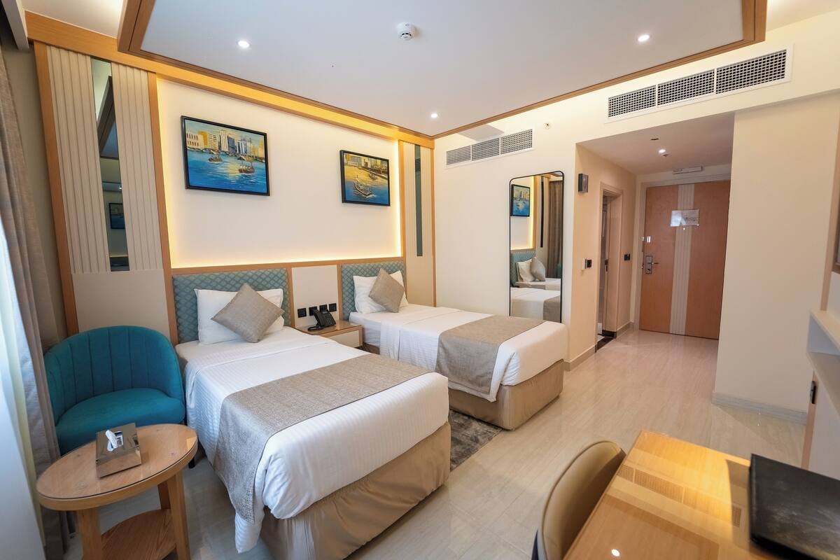 A hotel room with two beds, a small table, a chair, and a desk. There are paintings on the walls and a mirror above the bed.