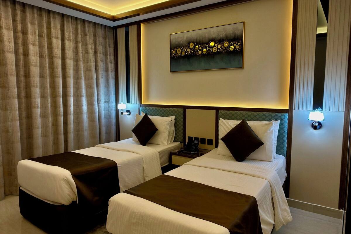 A modern hotel room with two beds, a large painting on the wall, and a ceiling light fixture.