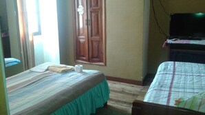 Una habitació amb dos llits, una porta de fusta, una televisió sobre un suport i una finestra amb cortines.