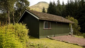 A small, green wooden cabin with a sloped roof, surrounded by dense forest and a hill in the background. - Beautiful 4 star, 2 bedroom chalet with mountain views. A true Highland landscape all around (Crianlarich)
