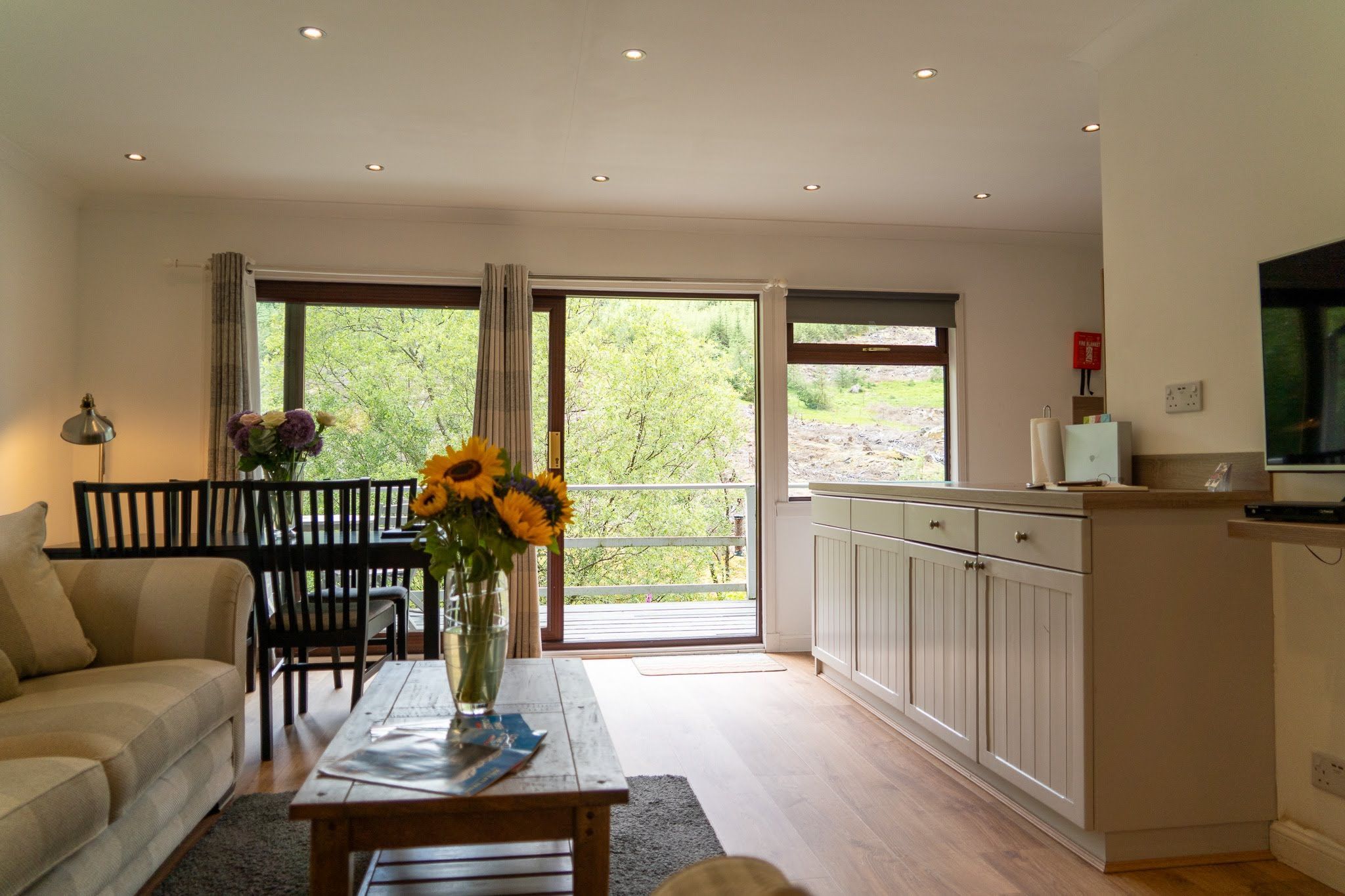 A living room with a sofa, a coffee table with a vase of flowers, a dining area with chairs and a table, and a kitchen with cabinets and a television.