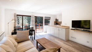 A modern living room with a sofa, a wooden coffee table, and a TV. There is a kitchen area with cabinets and a microwave. Large windows provide natural light and a view of trees. - Beautiful 4 star, 2 bedroom chalet with mountain views. A true Highland landscape all around (Crianlarich)