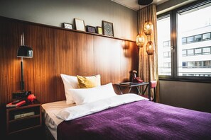 Une chambre d’hôtel avec une tête de lit en bois, un lit avec une literie blanche et violette, une table de chevet avec une lampe et une fenêtre donnant sur des immeubles.