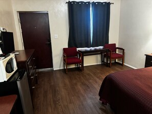 Quarto de hotel pequeno com piso de madeira, uma escrivaninha com duas cadeiras, uma cama, televisão e micro-ondas.