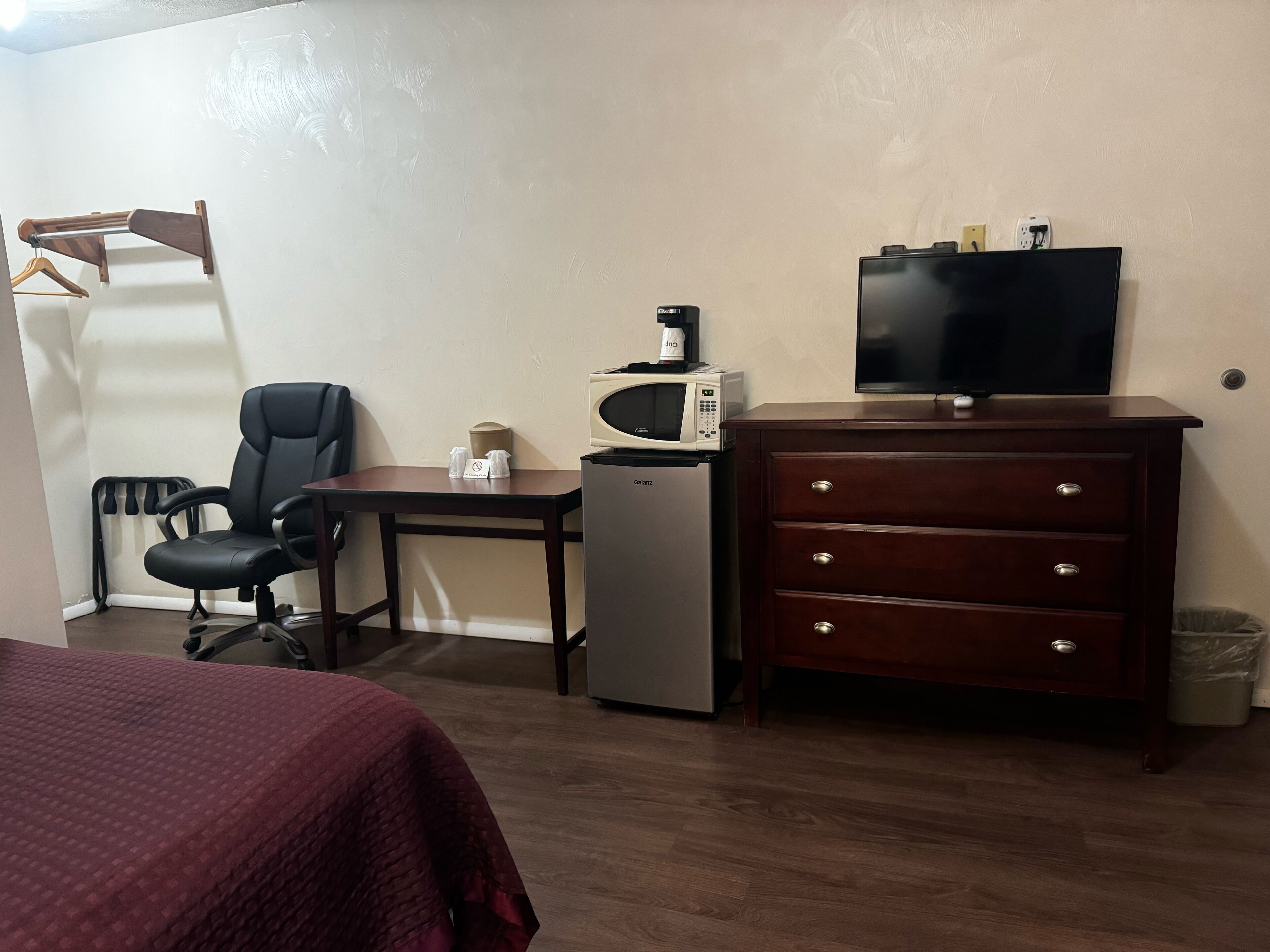 A hotel room with a bed, a desk with a chair, a microwave, a small refrigerator, a dresser, and a television.