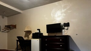 Ett kompakt hotellrum med ett skrivbord, ett litet kylskåp, en lampa, en tv och en byrå.