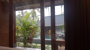 A view through a window with a wooden frame, revealing a pool area with a tiled roof, surrounded by various plants and palm trees. - Buda Cottage Ubud (Ubud)
