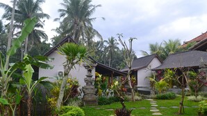 A tropical garden with a stone pathway leading to a white building with a tiled roof. - Buda Cottage Ubud (Ubud)