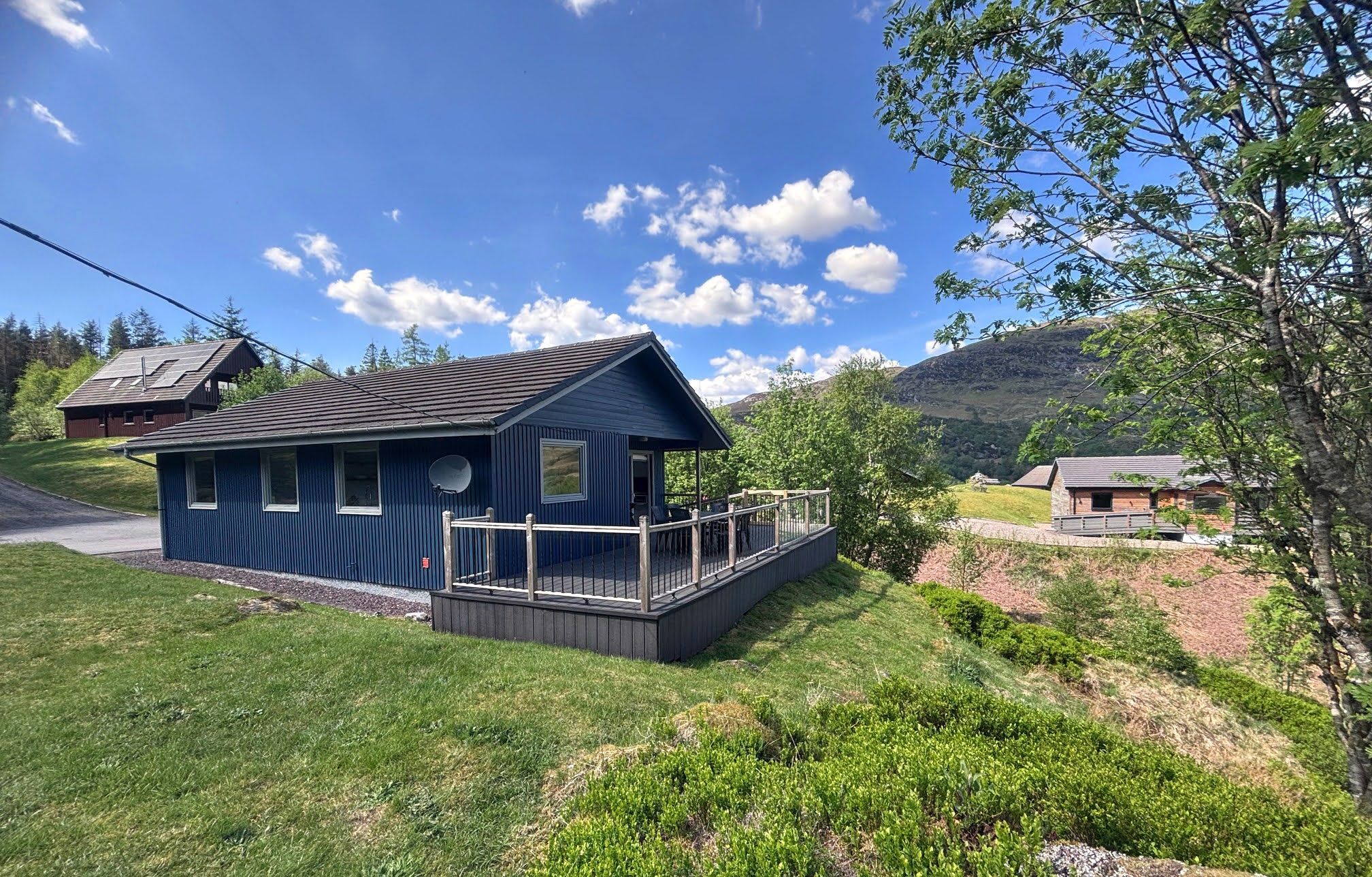 A small blue cabin with a deck, surrounded by greenery and hills.