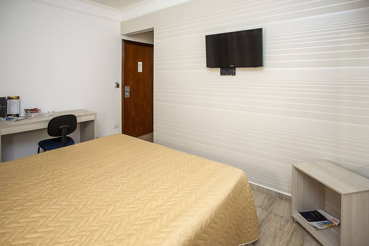 A hotel room with a bed, a desk, a chair, a television mounted on the wall, and a door.