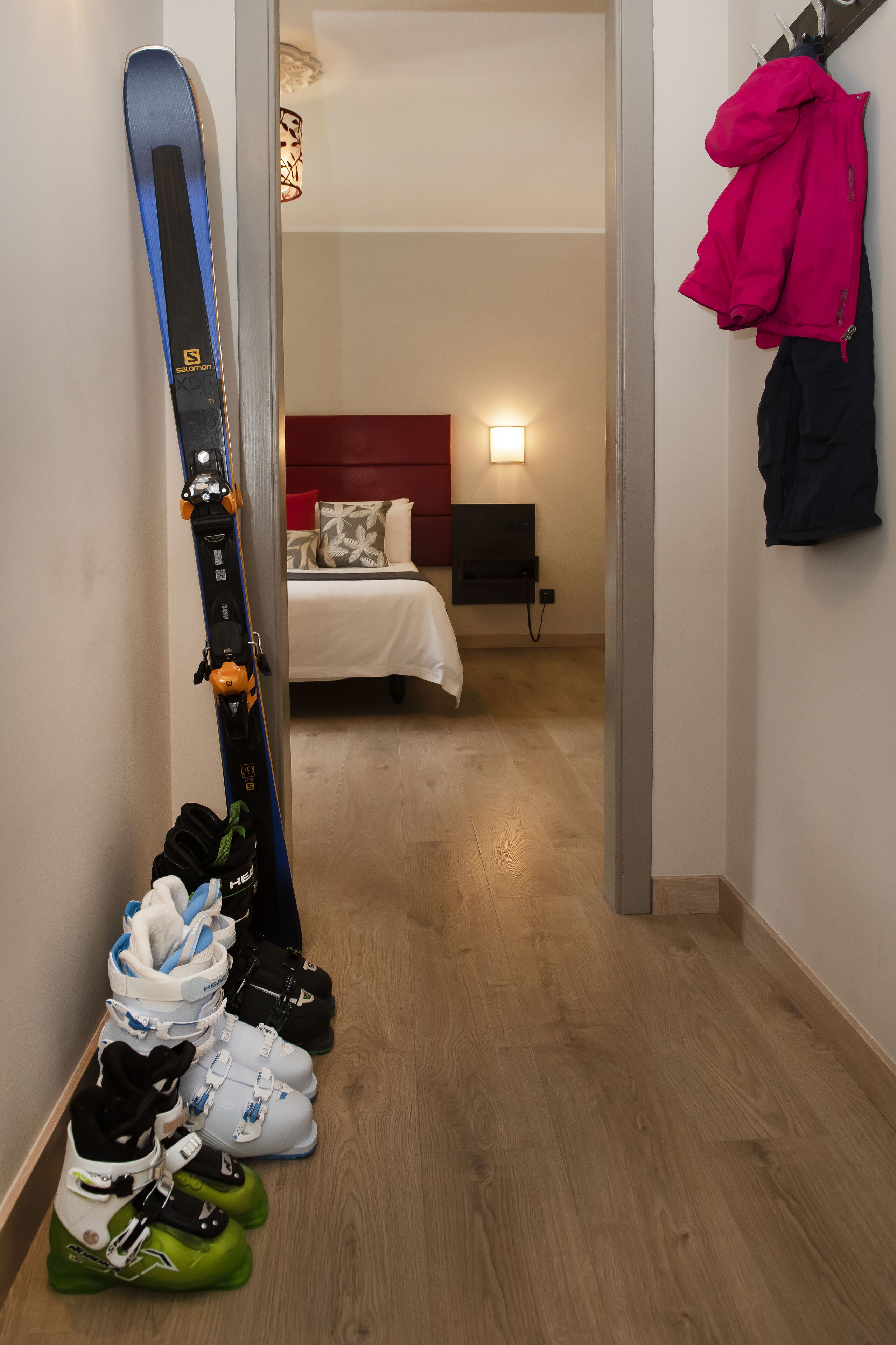 Un pasillo con esquíes y botas de esquí apoyados en la pared, una cama con cabecera roja y un perchero con chaquetas.