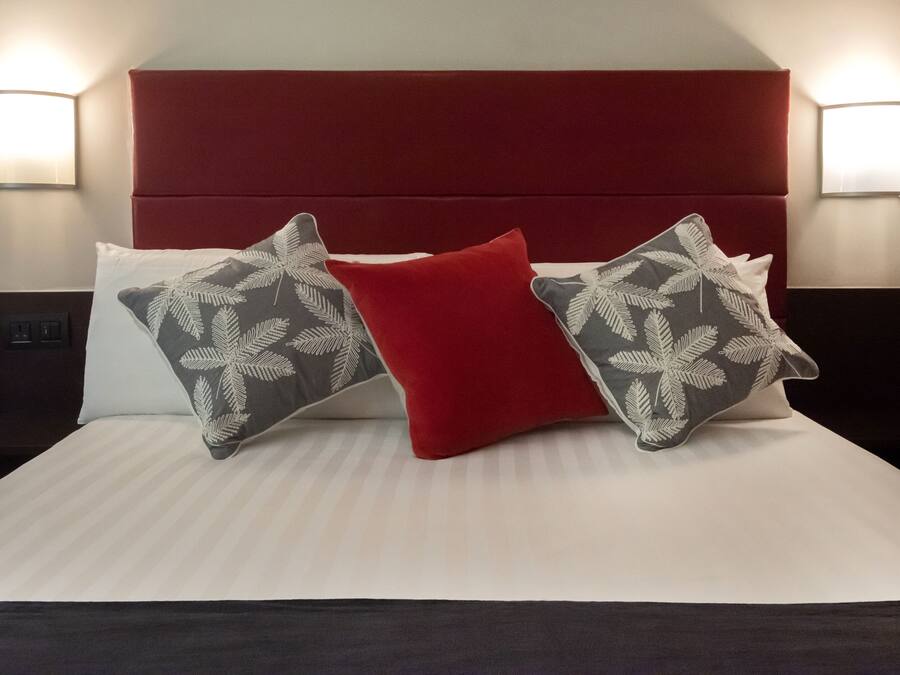 Una cama bien tendida con un cabecero rojo, dos almohadas estampadas y dos lámparas fijadas a la pared.