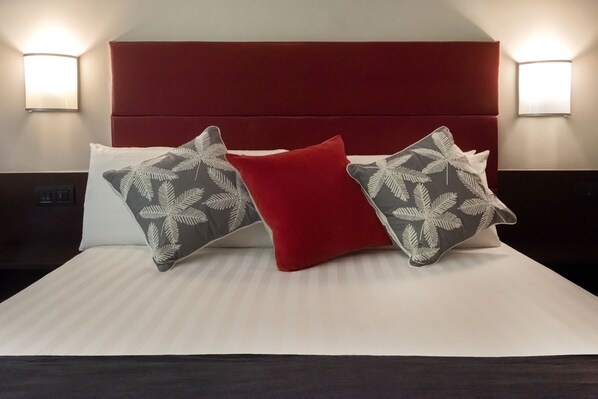 Una cama bien tendida con un cabecero rojo, dos almohadas estampadas y dos lámparas fijadas a la pared.