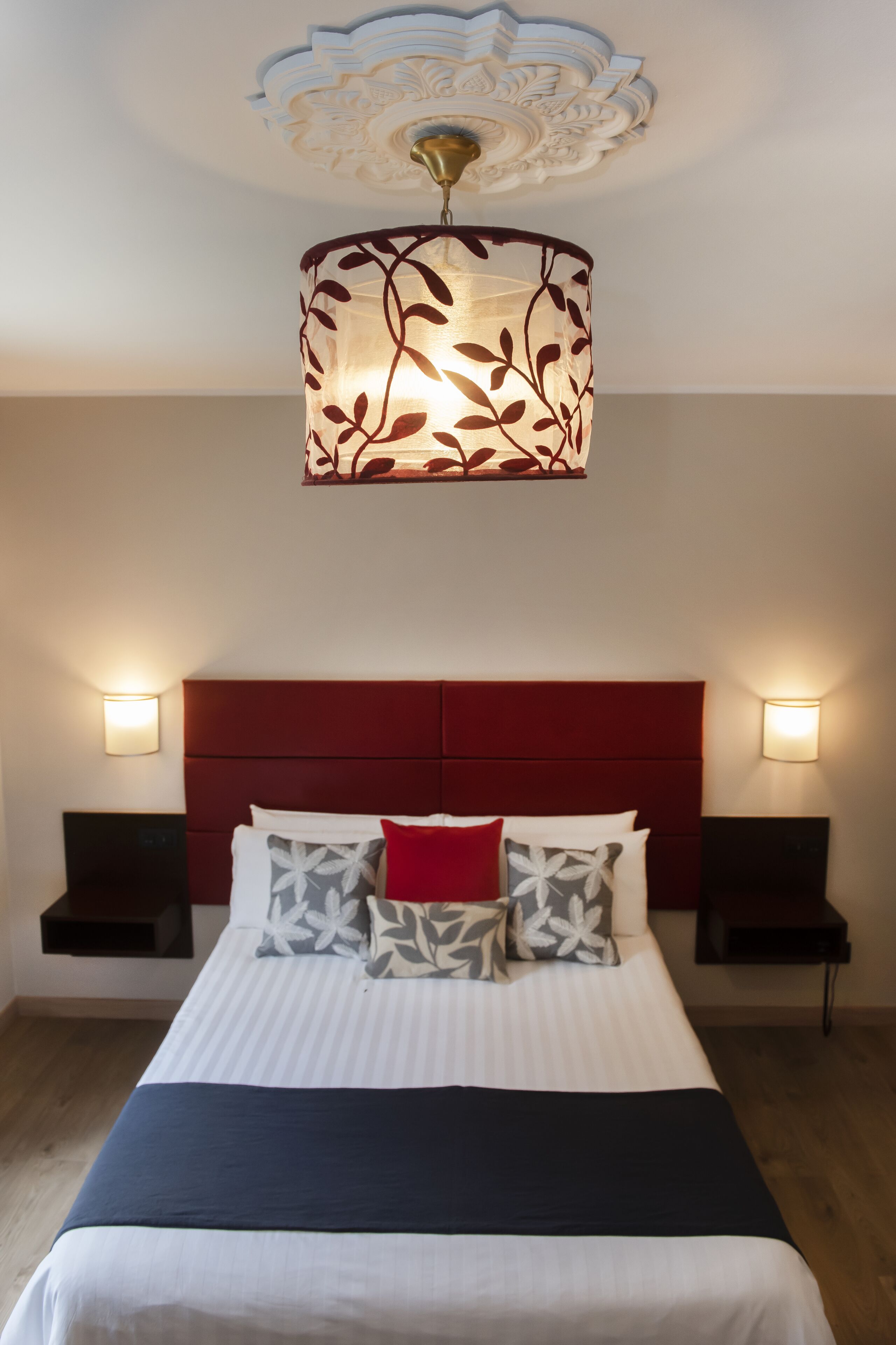 Een hotelkamer met een bed, een rode hoofdbord, twee nachtkastjes en een decoratieve plafondlamp.