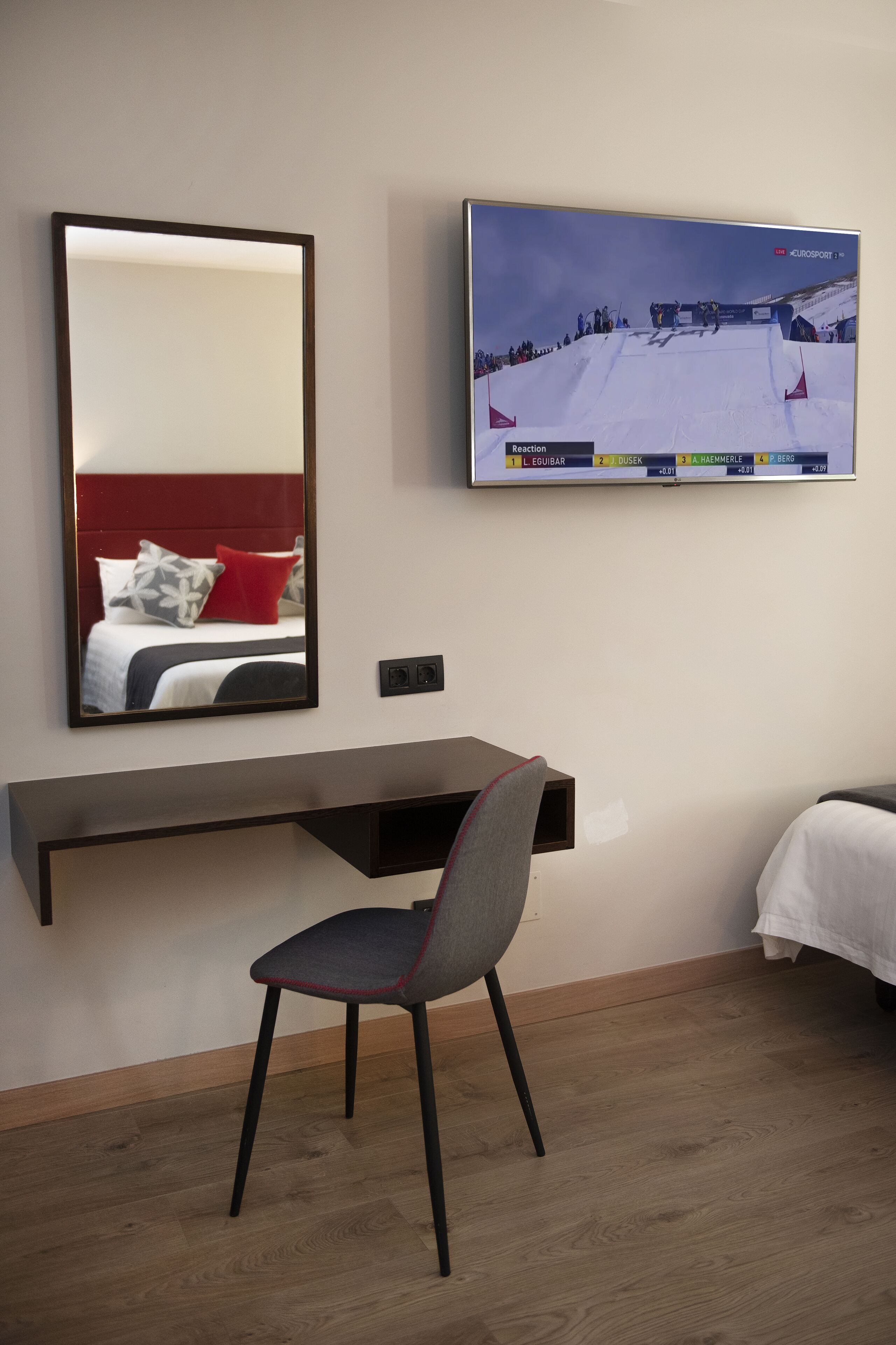 Habitación de hotel con cama, escritorio, silla, espejo grande y televisor mostrando un evento de esquí.