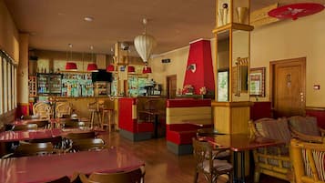 Un restaurante con mesas y sillas de madera, una zona de bar con estantes llenos de botellas y una pared roja decorativa.