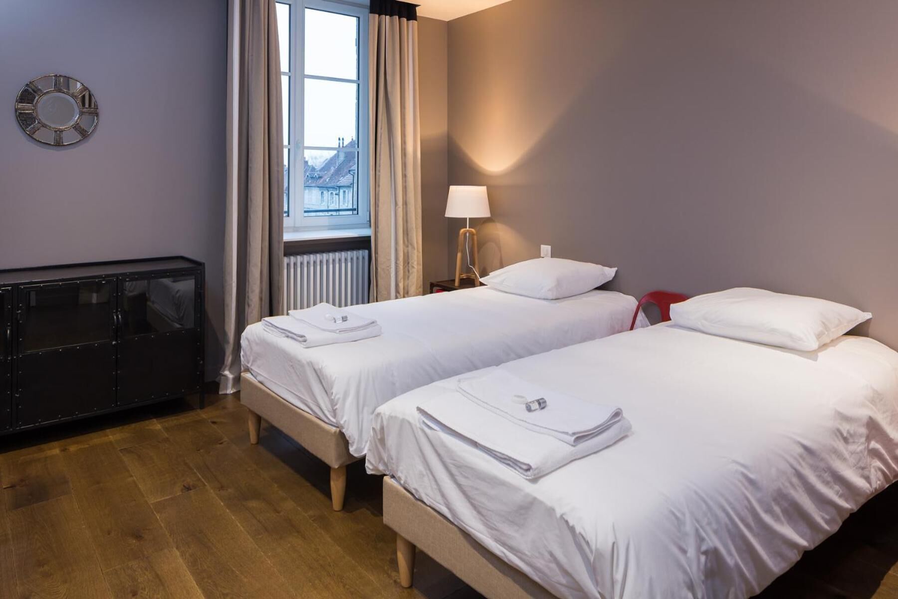 Une chambre d’hôtel avec deux lits, un radiateur, une horloge au mur et une fenêtre avec des rideaux.