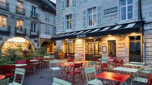 An outdoor cafe area with tables and chairs, a stone building with a sign for 'DOCKS BRASSERIE' in the background, and a small garden area. - Apparthôtels de Val-Perrière (Arbois)