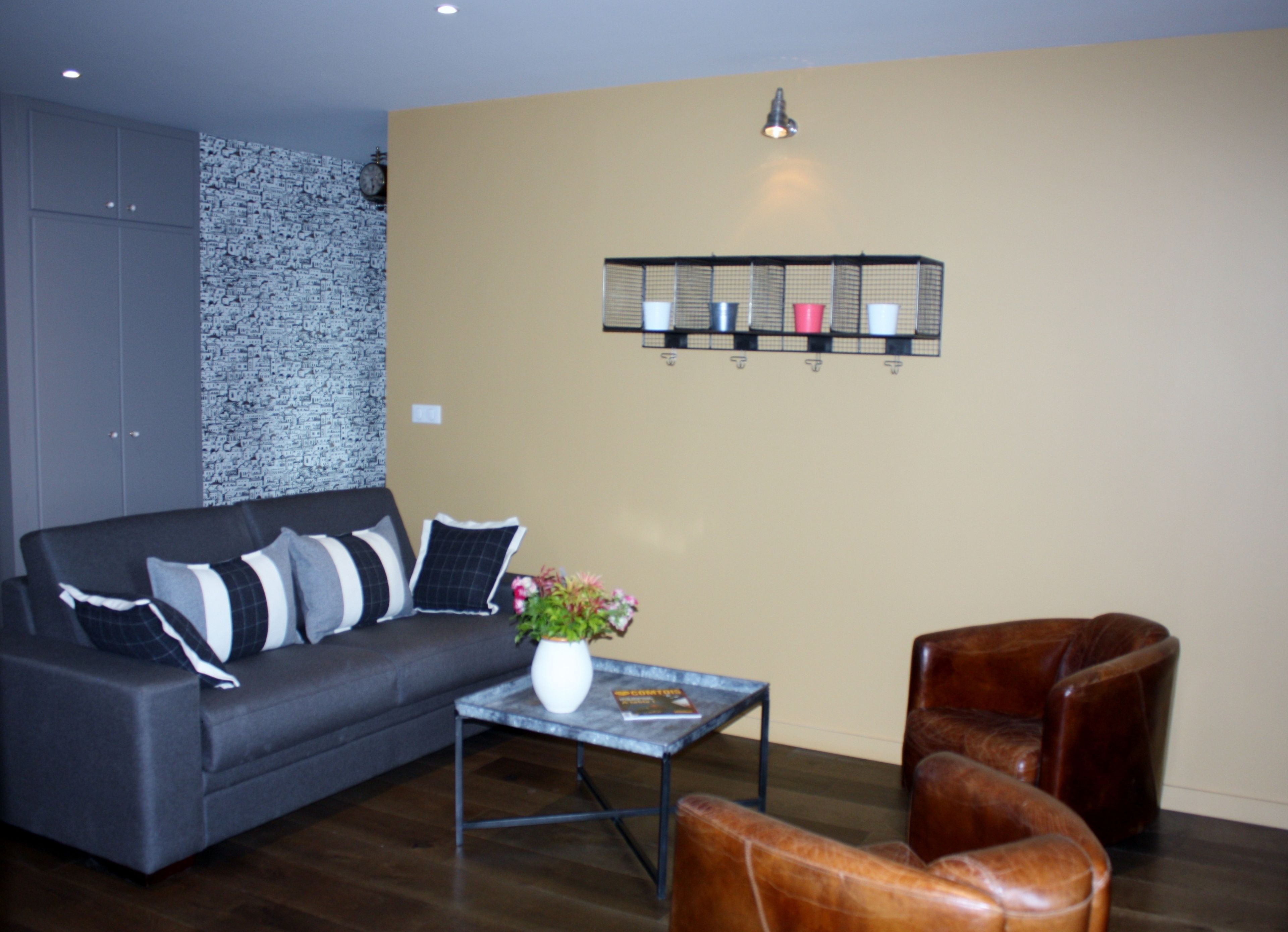 A living room with a grey sofa, a coffee table with a vase, a brown leather chair, and a wall-mounted shelf with cups.