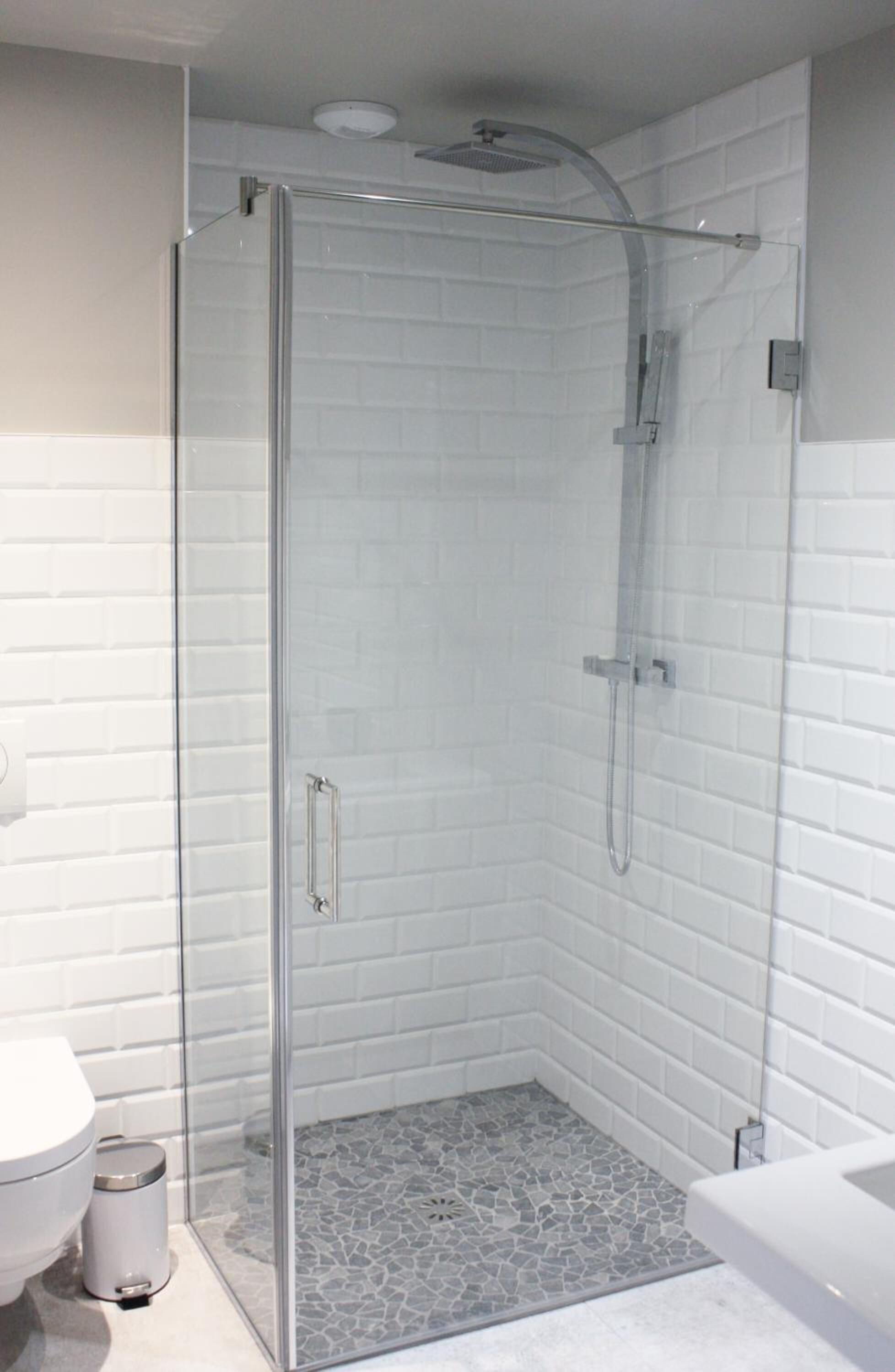 A modern bathroom with a glass-enclosed shower, white tiled walls, and a small trash bin.