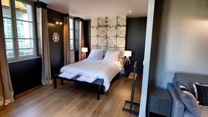 A bedroom with a bed, bedside lamps, a nightstand, and a view of the outside through a window. - Apparthôtels de Val-Perrière (Arbois)
