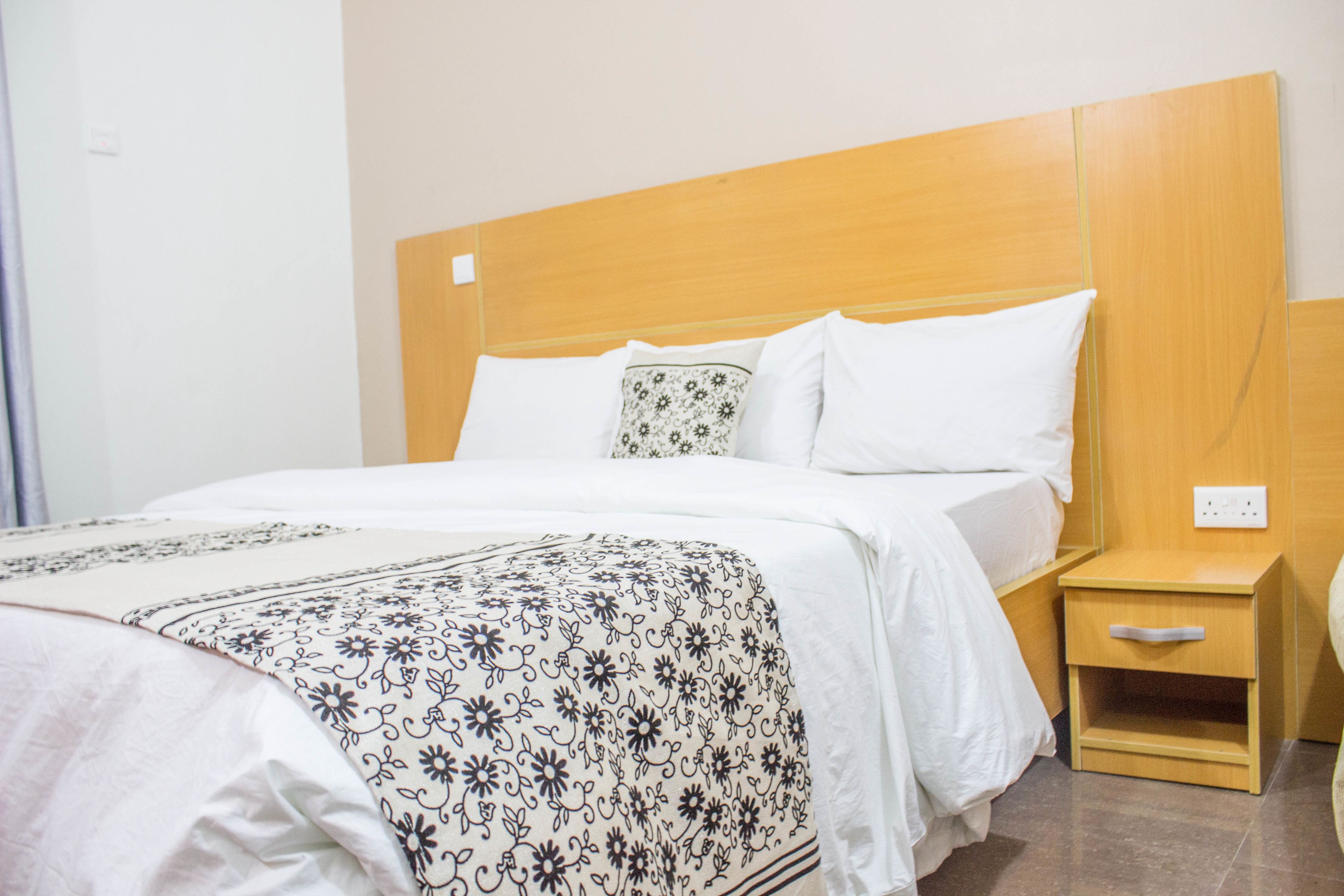 Een hotelkamer met een houten hoofdeinde, een bed met wit beddengoed, een bed met een gestreept plaid en een houten nachtkastje met een lade.