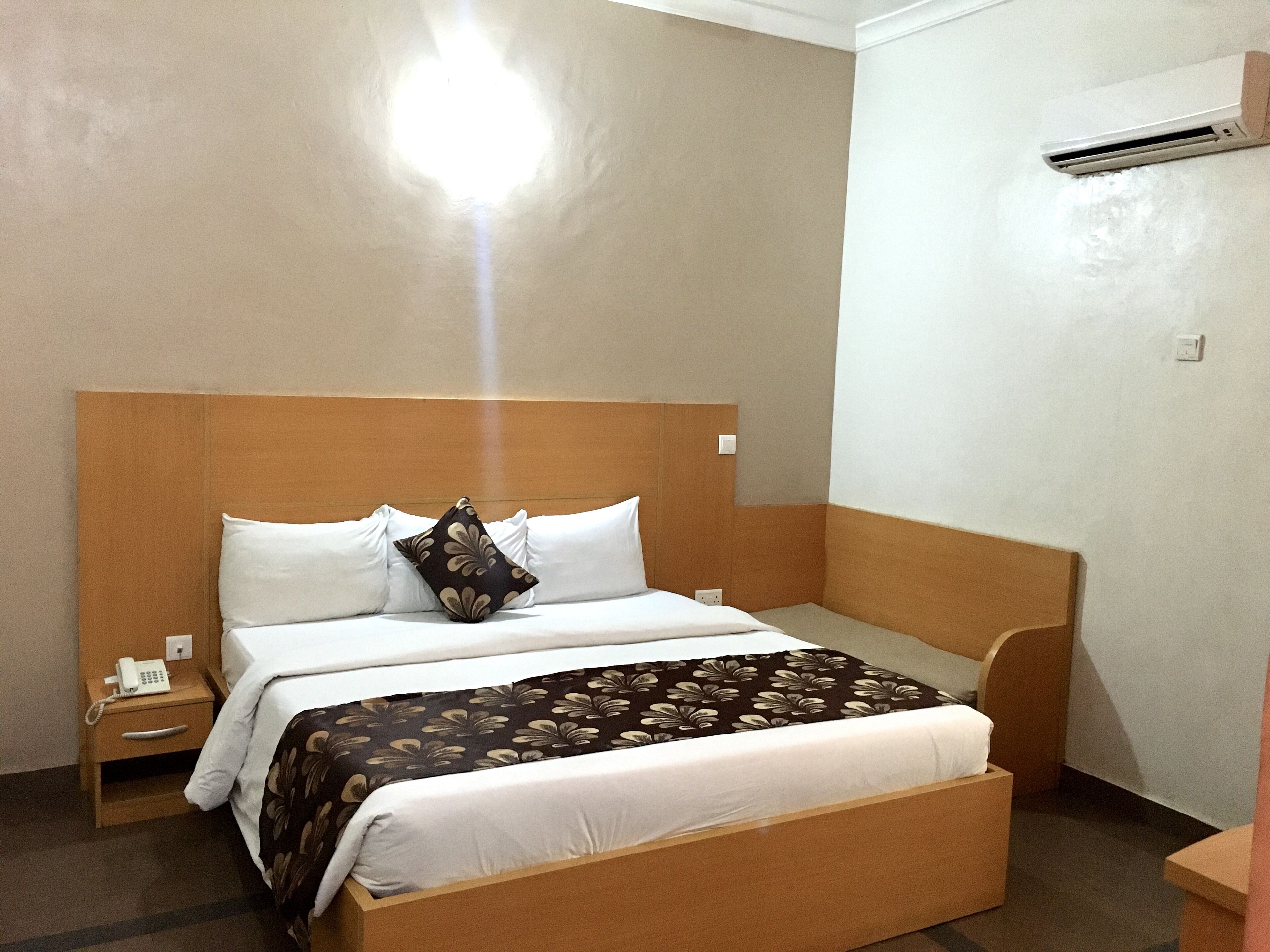 Habitació d'hotel amb un capçal de llit de fusta, un llit amb roba de llit blanca, una manta decorativa i una taula de nit amb un telèfon, i una unitat de climatització a la paret.