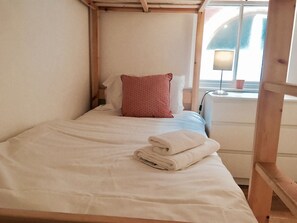 Una cama individual con ropa blanca y una almohada roja estampada, una toalla blanca doblada y una mesita de noche con una lámpara y una taza.